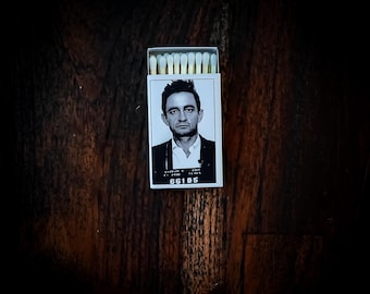 May include: A matchbox featuring a black and white image of a man in a mugshot. The matchbox has a white background with the man's image and the number "66185" printed on it. The match heads are visible at the top.