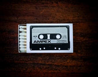 May include: A rectangular matchbox featuring a black and white graphic of an Ampex C-60 professional audio cassette tape on the cover. The box is partially open, revealing a row of wooden matches with white tips inside the cardboard sleeve.