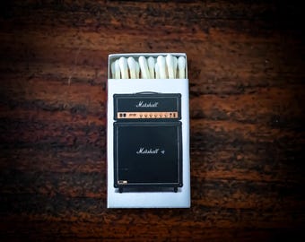 May include: A rectangular matchbox with a white exterior featuring a graphic illustration of a black Marshall guitar amplifier stack. The box is partially open at the top, revealing a row of wooden matches with white tips arranged inside the container.