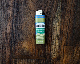 May include: A green and blue lighter with the word "pickles" and "Kosher Dill Spears" printed on it. The lighter also has an image of pickles and the text "25 FL OZ". The lighter is on a dark brown wooden surface.