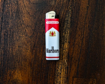 May include: A red, white, and gold Marlboro lighter. The lighter has the Marlboro logo and text on a white background, with red accents. The lighter is on a dark wood surface.