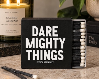May include: A black matchbox with white text that reads DARE MIGHTY THINGS (TEDDY ROOSEVELT) containing matches with black sticks and white tips. Two loose matches with black sticks and white tips lie in front of the box on a stone surface.