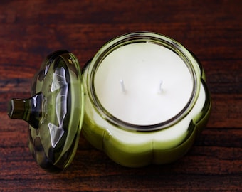 May include: A pumpkin-shaped olive green glass jar candle with a matching lid placed to the side. The candle features smooth white wax and two visible wicks. The glass container has a ribbed, segmented exterior design resembling a gourd.
