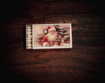 May include: A matchbox with a festive Christmas theme. The box features a vintage illustration of Santa Claus in a red suit, holding greenery. The match heads are white.
