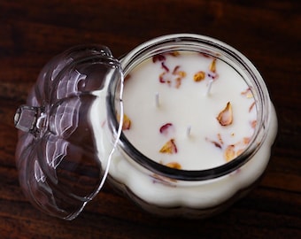 May include: A clear glass pumpkin-shaped jar containing a white candle with three wicks. The surface of the wax is decorated with scattered dried flower petals. The matching glass lid is placed to the side of the open jar, revealing the interior.