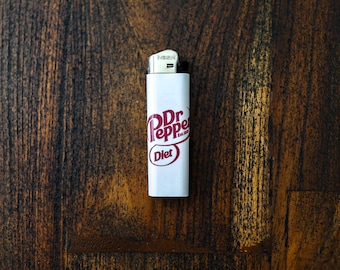 May include: A white lighter with the "Dr Pepper Diet" logo in red. The lighter is set against a dark wood grain background. The lighter is a standard size.
