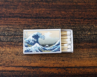 May include: A rectangular matchbox featuring a print of The Great Wave off Kanagawa on the cover. The box is partially open, revealing a row of wooden matches with white tips arranged inside the white cardboard casing.