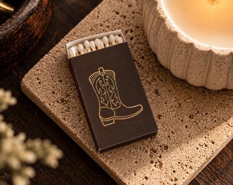 May include: A rectangular dark brown matchbox featuring a gold-colored line drawing of a cowboy boot on the front. The box is partially open, revealing a row of wooden matches with white tips inside the cardboard container.