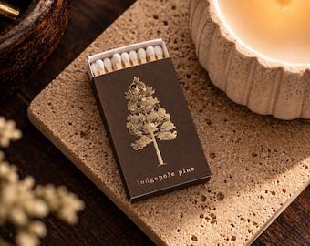 May include: A rectangular matchbox with a dark brown cover featuring a gold foil illustration of a pine tree and the text "lodgepole pine" printed in gold. The box is partially open, revealing a row of wooden matches with white tips.
