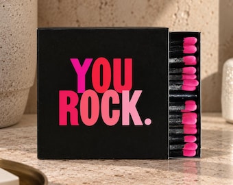 May include: A square black matchbox featuring the text YOU ROCK in bold pink and light pink lettering. The box is partially open, revealing a row of matches with black wooden sticks and bright pink match heads arranged in a vertical line.