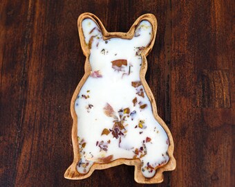 May include: A dog-shaped wooden bowl filled with white wax and embedded with dried flower petals. The bowl features a smooth, light-brown wood grain finish and is molded into the silhouette of a dog with pointed ears and a tail.