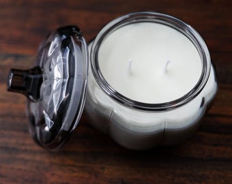 May include: A pumpkin-shaped glass candle jar with a matching lid placed to the side. The jar contains white wax and features two visible wicks. The glass is dark and translucent, with a ribbed exterior design that mimics a pumpkin's shape.