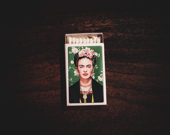 May include: A matchbox featuring a portrait of Frida Kahlo with a floral crown and jewelry. The background is green with white flowers. The matchbox is open, revealing white matchsticks.