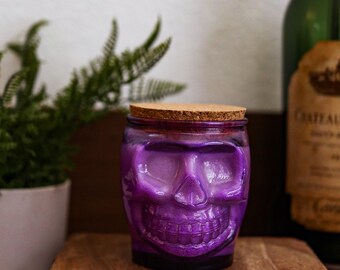 May include: A purple glass jar shaped like a human skull with a cork lid. The glass is translucent and features a molded facial structure including eye sockets, a nose cavity, and teeth. The jar is cylindrical with a flat base and a circular top.