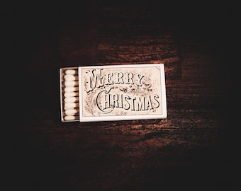 May include: A matchbox with the words "Merry Christmas" in an ornate font. The matchbox is open, revealing a row of white-tipped matches. The background is a dark, textured wooden surface.