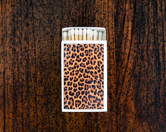 May include: A matchbox with a leopard print design. The box is white with a rectangular leopard print pattern in shades of brown and black. The matches are visible at the top, with white tips and wooden stems. The background is a dark brown wooden surface.