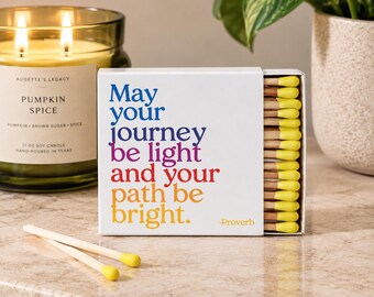 May include: A white rectangular matchbox featuring the text "May your journey be light and your path be bright. -Proverb" in blue, purple, red, and orange. The box contains wooden matches with bright yellow tips, with two matches lying outside.