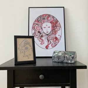 May include: A black framed print of a red and white illustration of a woman's face with a circle around it. A smaller black framed print of a black and white illustration of a person is to the left of the larger print. A small gray and black box is in front of the prints on a black table.