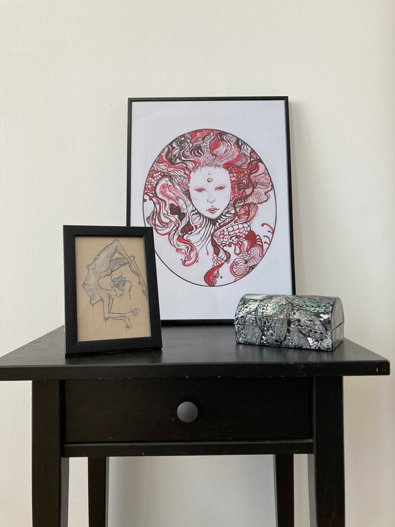 May include: A black framed print of a red and white illustration of a woman's face with a circle around it. A smaller black framed print of a black and white illustration of a person is to the left of the larger print. A small gray and black box is in front of the prints on a black table.
