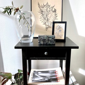 May include: A black side table with a drawer, a small black picture frame, a black and white drawing of a seashell, a clear glass vase with an olive branch, a small green and black box, and a stack of books with the titles "Quite Lovely" and "Ren Hang".