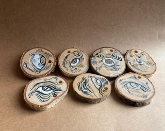May include: Seven wooden ornaments featuring black and white ink drawings of eyes. Each ornament is round and has a hole drilled through the top for hanging.