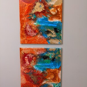 May include: Two abstract paintings on canvas with a mix of orange, red, blue, green, and gold paint. The paintings are displayed side-by-side on a wall.