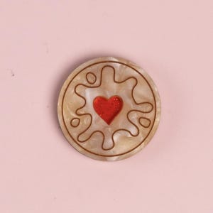 May include: A circular brooch with a cream-coloured, marbled effect. It features a red, glittery heart in the centre, surrounded by a brown, stylised design. The brooch is set against a soft pink background.