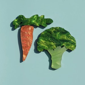 Puede incluir: Adornos decorativos con forma de zanahoria y brócoli. La zanahoria es naranja con hojas verdes. El brócoli es verde con un tallo verde claro, sobre fondo azul claro.