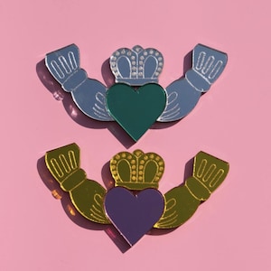 Puede incluir: Dos símbolos de Claddagh, uno en azul y verde, el otro en oro y morado, se muestran sobre un fondo rosa. Cada símbolo presenta un corazón, una corona y manos, que representan el amor, la lealtad y la amistad. Los símbolos son reflectantes.