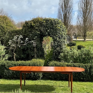 May include: An oval wooden table with a warm, reddish-brown hue, set on a lawn. The table has four slender legs and a smooth surface. The design suggests a mid-century modern style. The table's length is approximately 200 cm.