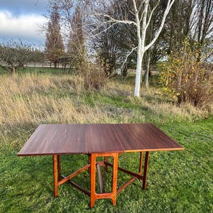 Puede incluir: Una mesa rectangular de madera marrón oscuro con diseño plegable. La mesa tiene un rico tono marrón rojizo y está sostenida por cuatro patas y una estructura de soporte central. La mesa está colocada al aire libre sobre una zona de césped. Mide unos 120 cm.