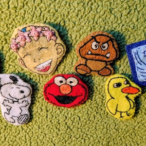 May include: A collection of seven embroidered felt patches featuring various characters and a rectangular blue patch with the embroidered text "this too shall pass". The designs include a green creature, a smiling face with pink flowers, a brown character, a white dog, a red character, and a yellow duck.