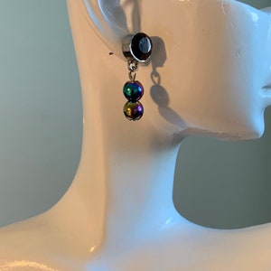 May include: A silver earring featuring a dark red gem and two iridescent spherical beads. The beads display a spectrum of colours, including blue, green, and gold. The earring is displayed on a white mannequin head.