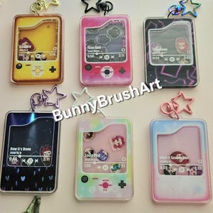 May include: A collection of six colorful acrylic keychains, each designed to resemble a retro music player. The keychains feature various song titles and artist names, with star-shaped charms and metal clasps. The keychains have a transparent design.