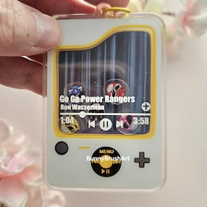 May include: A rectangular acrylic keychain featuring a digital music player interface with a yellow border. The screen displays Go Go Power Rangers by Ron Wasserman, a progress bar, and four Power Ranger helmet icons. A black menu button is below.