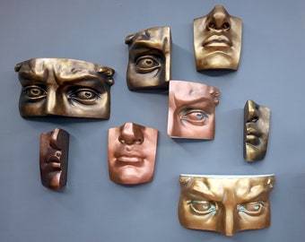May include: A collection of eight decorative wall sculptures featuring different human faces. The sculptures are made of a metallic material with a bronze finish. The faces are all unique and expressive, with different features and expressions.