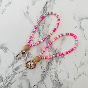 May include: Two colorful wrist keychains. Each keychain features a loop made of small, flat, colorful beads in shades of pink, blue, yellow, and white. A wooden bead and a metal clasp complete the design. The keychains are displayed on a white marble surface.