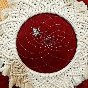 Peut inclure: Un attrape-rêves en macramé blanc avec un motif de toile d'araignée perlée et un accent d'araignée en bijoux. L'attrape-rêves est suspendu sur un fond de velours rouge.
