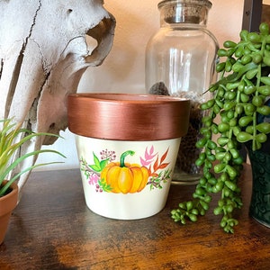 May include: A white ceramic planter with a copper rim, featuring a hand-painted design of a pumpkin with fall foliage and flowers. The planter is sitting on a wooden surface.