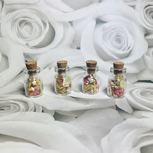 May include: Four small glass bottles with cork stoppers filled with dried flower petals in shades of pink, white, and yellow. The bottles are arranged in a row on a white background.