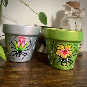 May include: Two painted terracotta flower pots with black spider and flower designs. One pot is silver with a pink rose and the other is green with a yellow hibiscus.