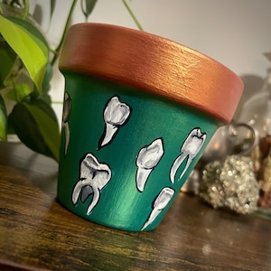 May include: A green terracotta planter with a copper-colored rim. The planter is decorated with white painted teeth.