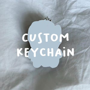 May include: A light blue, irregularly shaped keychain featuring the white text CUSTOM KEYCHAIN centred on its surface. The keychain includes a small silver metal ring attachment at the top, designed for securing keys or other small personal items.