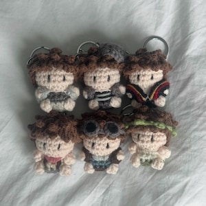 Puede incluir: Seis pequeños llaveros de ganchillo con figuras de pelo de lana marrón y piel beige claro. Cada figura viste ropa de diferentes colores, incluyendo detalles en gris, negro, rojo, amarillo y verde, unidos a anillas de metal plateado.