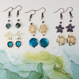 May include: Three pairs of dangle earrings with a sea-themed design. Each pair features a different charm: a turtle, a starfish, and a seashell. The earrings are made with blue and white beads and have a copper finish.