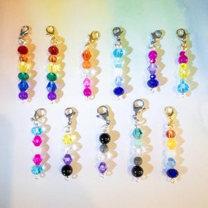May include: A collection of beaded charms with lobster claw clasps. The charms feature a variety of colorful beads, including red, orange, yellow, green, blue, and purple. Some charms have gold-toned clasps, while others have silver-toned clasps. The beads are arranged in vertical rows.