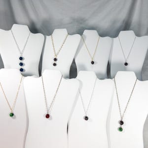 May include: A collection of necklaces displayed on white bust forms. Each necklace features a delicate chain and a pendant with one to three colorful, round beads. The beads are in various colors, including blue, red, green, and black.