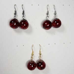 May include: Three pairs of dangle earrings. Two pairs have silver-colored hooks, and one pair has gold-colored hooks. Each earring features a round, red, glittery bead. The earrings are displayed against a white background.