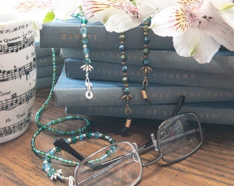 May include: Three beaded eyeglass chains with different colored beads and metal accents. The chains are laying on top of a stack of books with titles like "Hamlet", "Julius Caesar", "Shakespeare's Poems", and "The Taming of the Shrew".