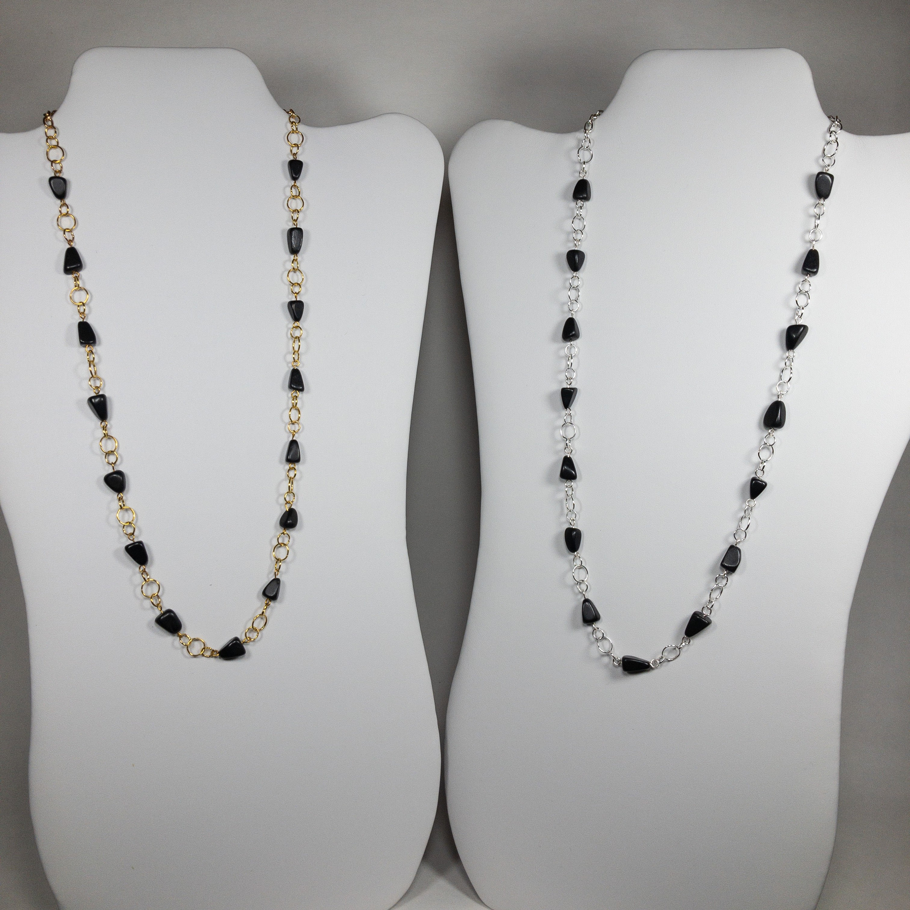 Puede incluir: Dos collares con cuentas triangulares negras en expositores. Un collar tiene una cadena dorada y el otro una cadena plateada.